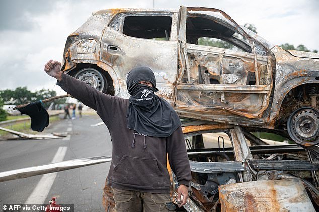 A masked bystander poses in front of burnt vehicles in a roadblock at the entrance to Ducos, France's Pacific territory of New Caledonia, on May 21, 2024