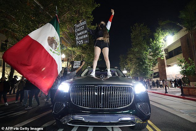 A woman stands on a car with a Mexican flag and a placard as protesters gather near the Federal Building in Santa Ana, California